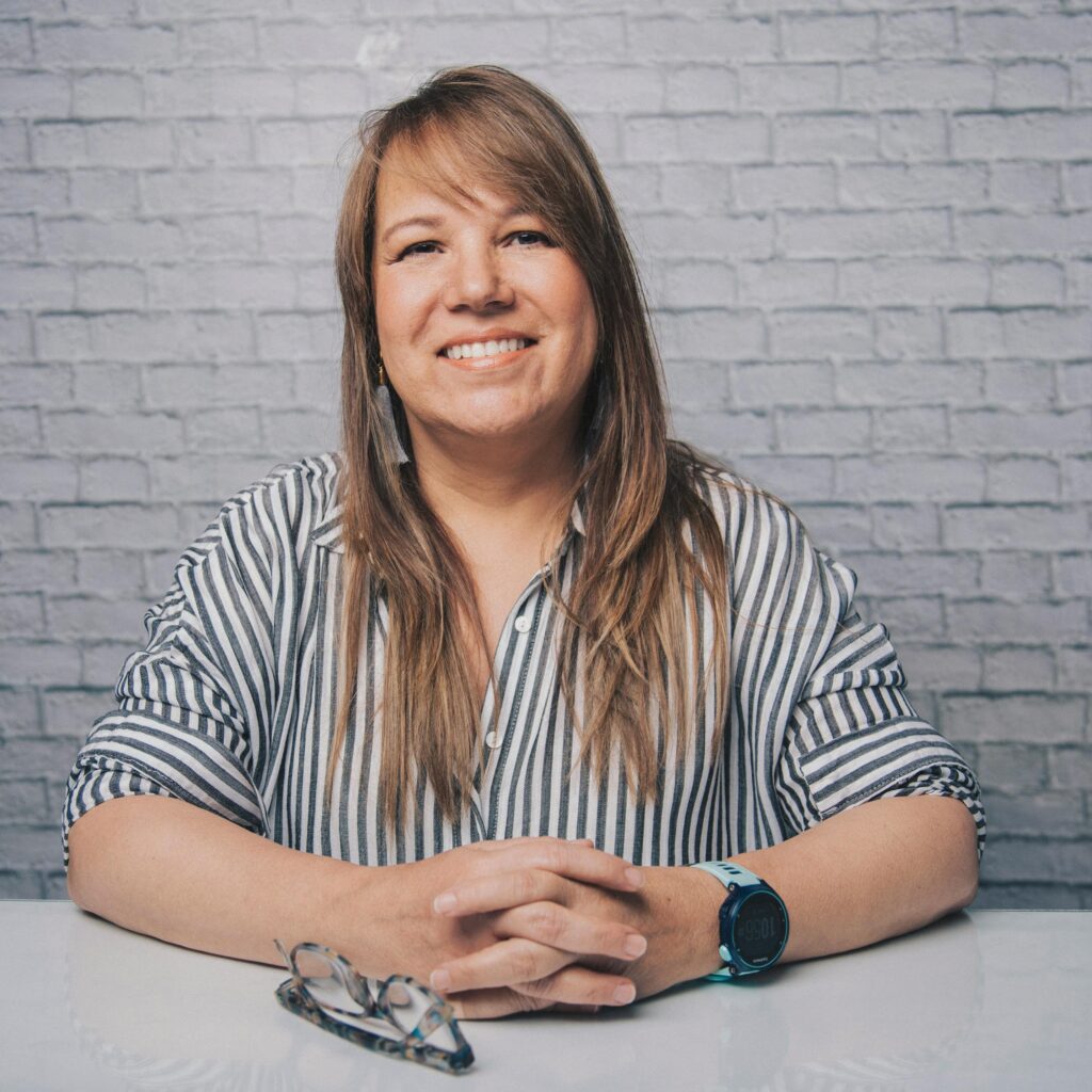 A cheerful woman with long hair and a striped blouse smiles warmly indoors against a brick wall.